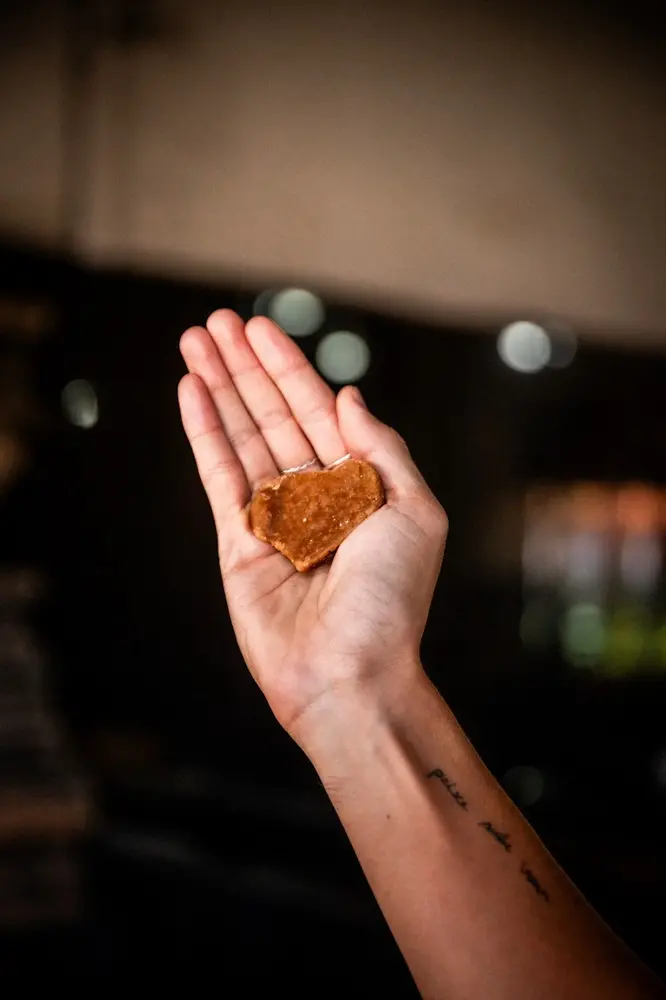 Na imagem, uma foto em close-up, com iluminação dramática e fundo muito escuro e desfocado, captura a mão estendida de uma pessoa segurando um pequeno pedaço de rapadura. O pedaço de rapadura é de cor caramelo-brilhante, tem uma forma irregular e repousa na palma aberta da mão. O braço e o pulso da pessoa estão visíveis. No antebraço, há uma tatuagem escrita à mão. O fundo escuro é interrompido por alguns pontos de luz circulares e desfocados (bokeh). O foco está totalmente no doce e na mão, destacando a textura e o formato do produto artesanal.