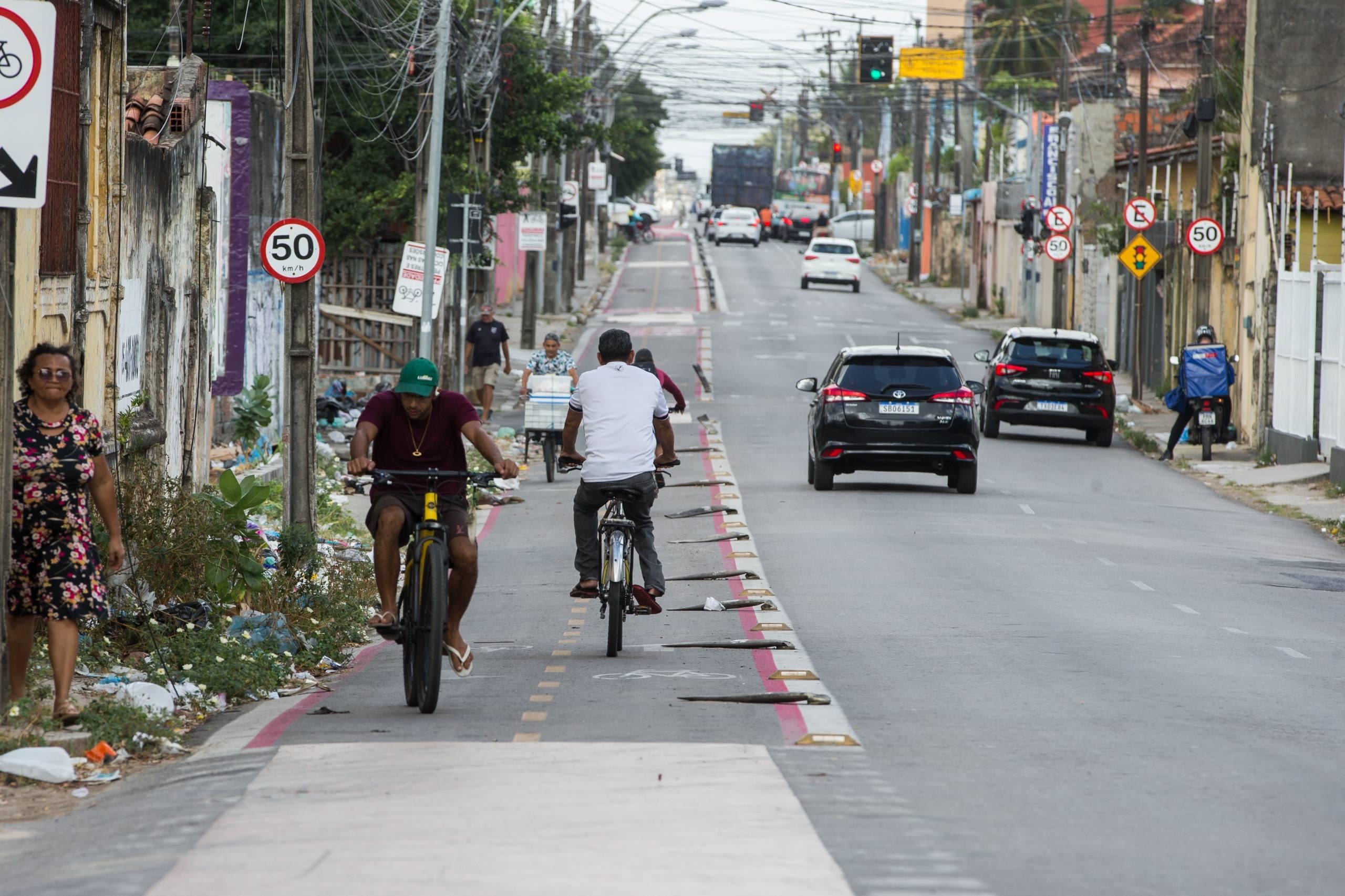 Vista de uma rua urbana movimentada, com ciclovia à esquerda. Dois ciclistas, um vestindo camiseta bordô e boné verde e outro de camiseta branca, pedalam na ciclovia de sentido único, separada da pista principal por tachões. Carros circulam na rua, e pedestres caminham na calçada lateral. Placas de limite de velocidade são visíveis.