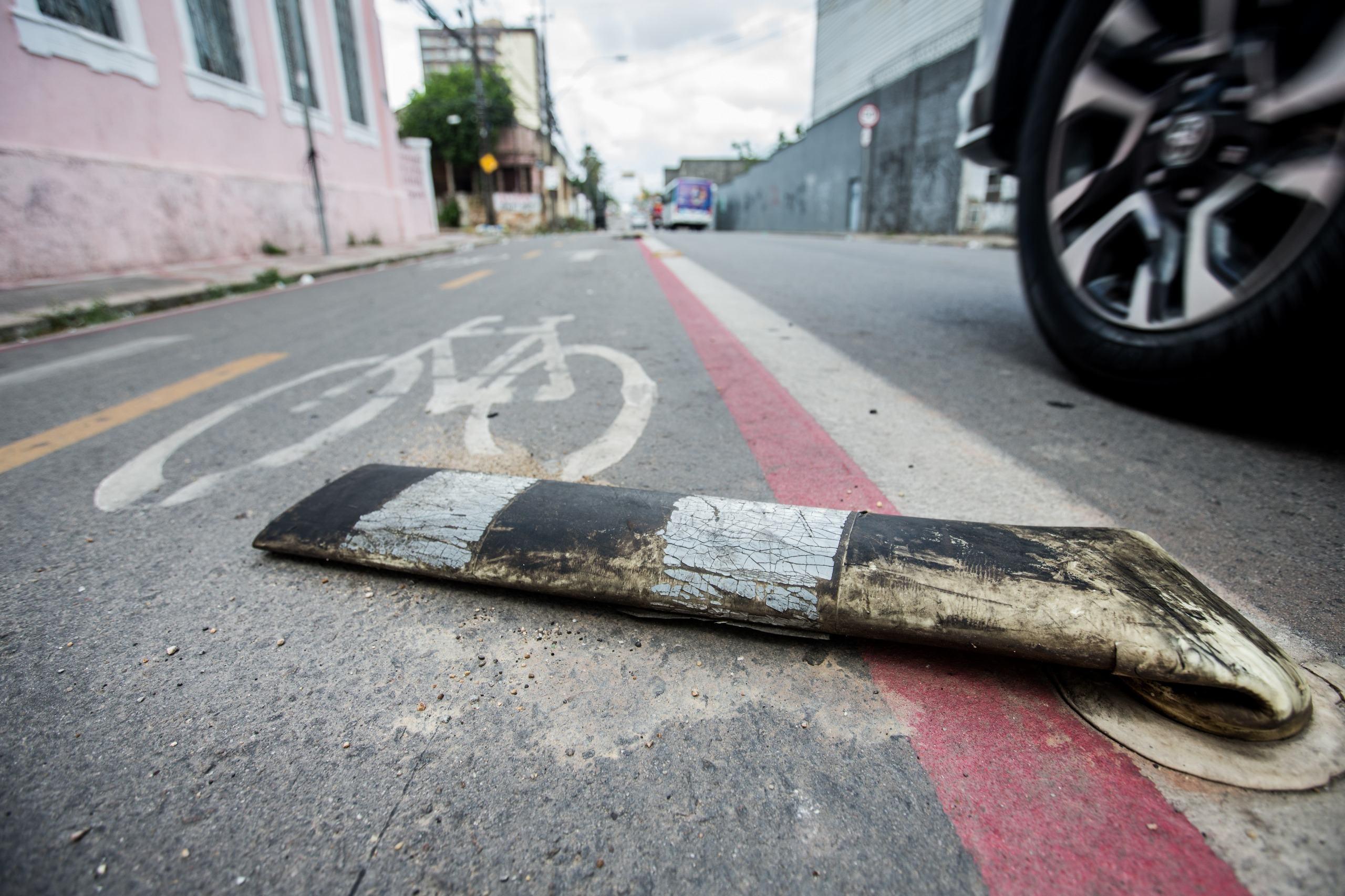 Poste delimitador de tráfego flexível, listrado em preto e branco e visivelmente danificado, caído sobre o asfalto. O poste se encontra dentro de uma ciclofaixa (indicada por um símbolo de bicicleta pintado e uma linha vermelha que a separa da pista de rodagem). A roda de um veículo ocupa o canto superior direito da imagem, enquanto edifícios antigos se alinham à esquerda, na distância.