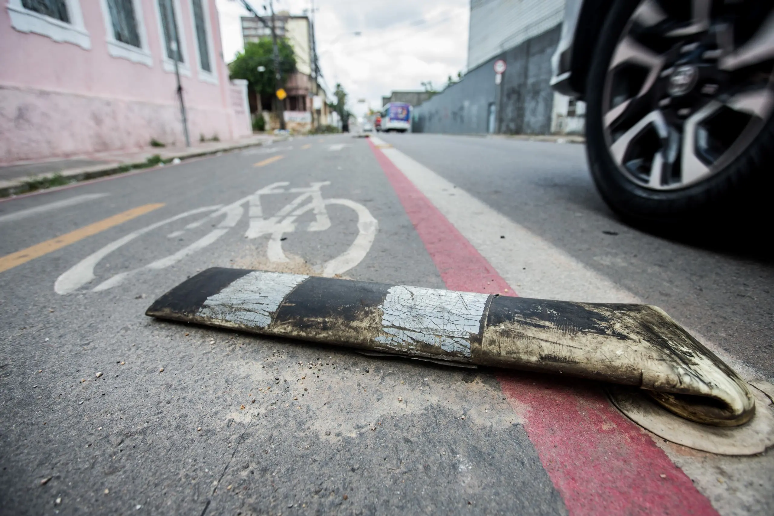 Poste delimitador de tráfego flexível, listrado em preto e branco e visivelmente danificado, caído sobre o asfalto. O poste se encontra dentro de uma ciclofaixa (indicada por um símbolo de bicicleta pintado e uma linha vermelha que a separa da pista de rodagem). A roda de um veículo ocupa o canto superior direito da imagem, enquanto edifícios antigos se alinham à esquerda, na distância.