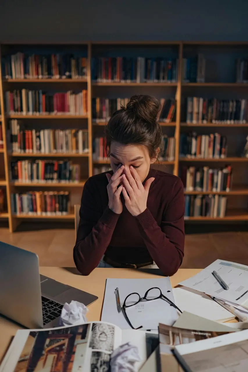 Pessoa sentada em uma biblioteca com as mãos no rosto, aparentando cansaço enquanto estuda. À sua frente há livros abertos, papéis espalhados, óculos, canetas e um notebook sobre a mesa. Estantes cheias de livros ocupam o fundo da cena.