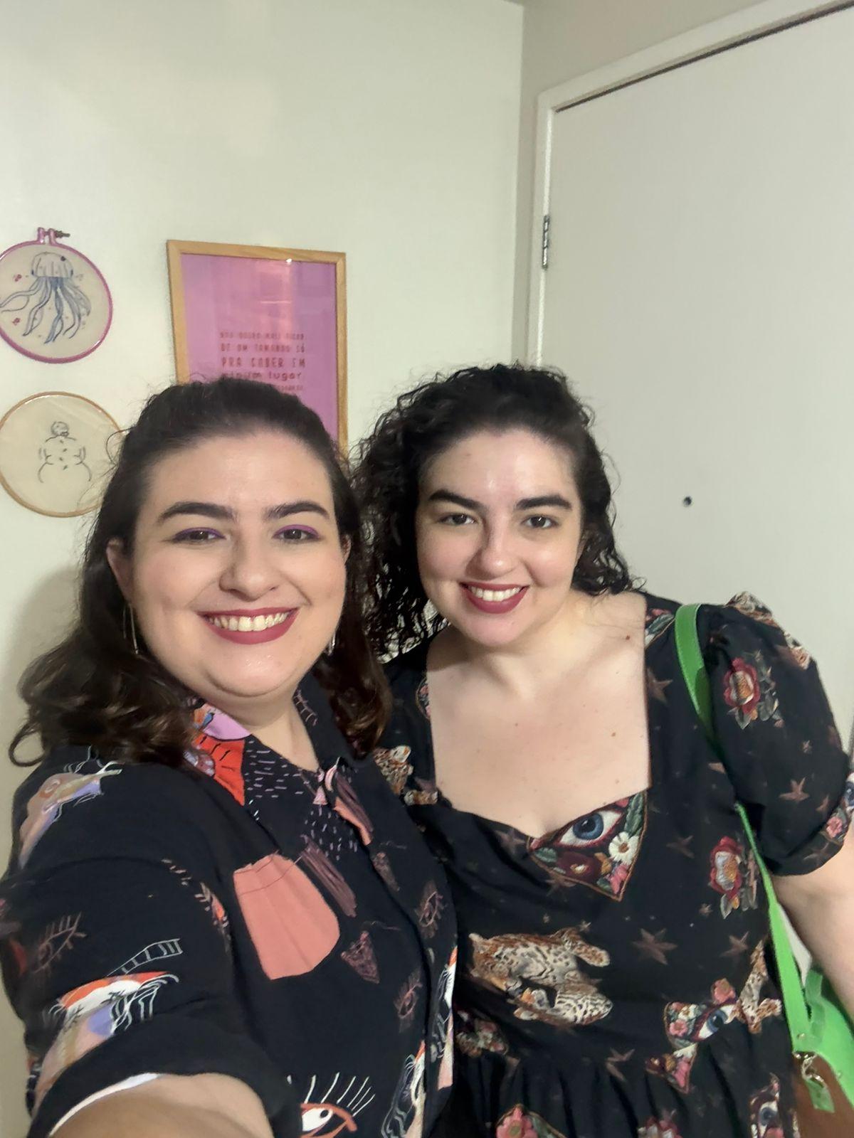 Duas mulheres sorrindo juntas na frente de uma parede com desenhos de peixes e uma moldura rosa, comemorando um momento especial.