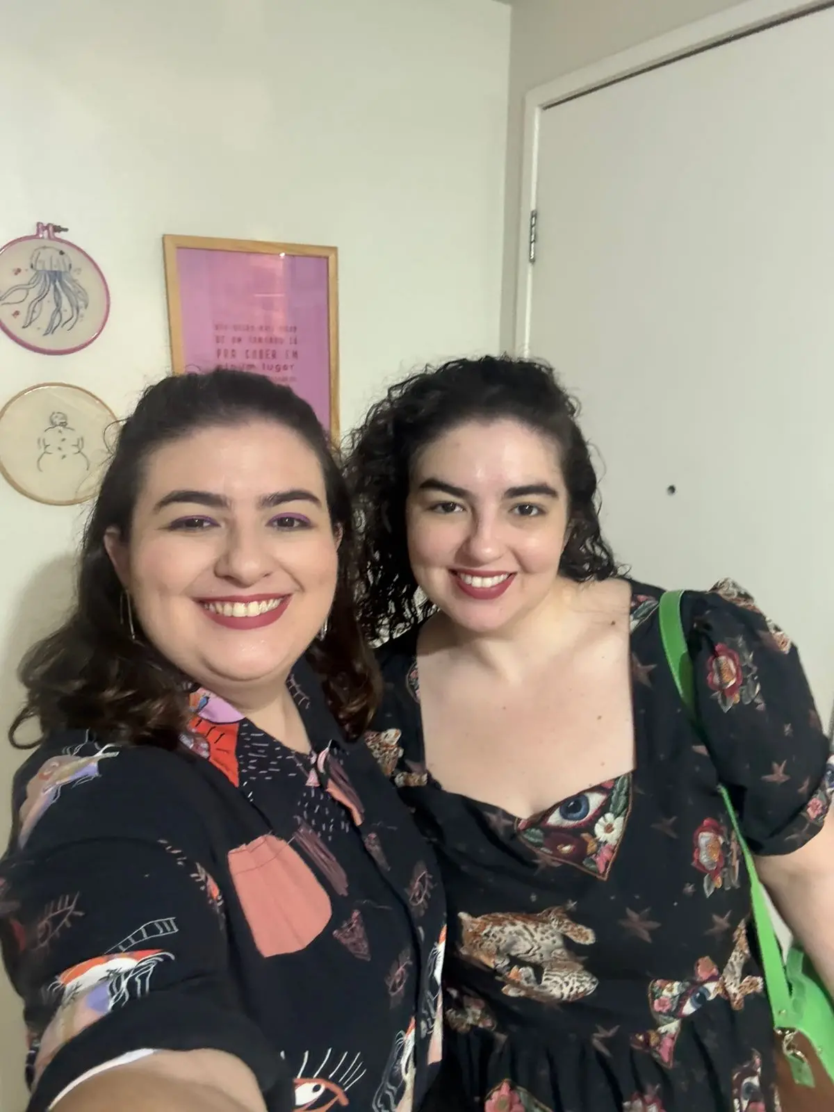 Duas mulheres sorrindo juntas na frente de uma parede com desenhos de peixes e uma moldura rosa, comemorando um momento especial.