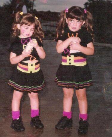 Duas meninas vestidas com roupas coloridas, cabelo de coque e maquiagem, posando juntas ao ar livre durante o pôr do sol, com maquiagem e cabelo bem arrumados.