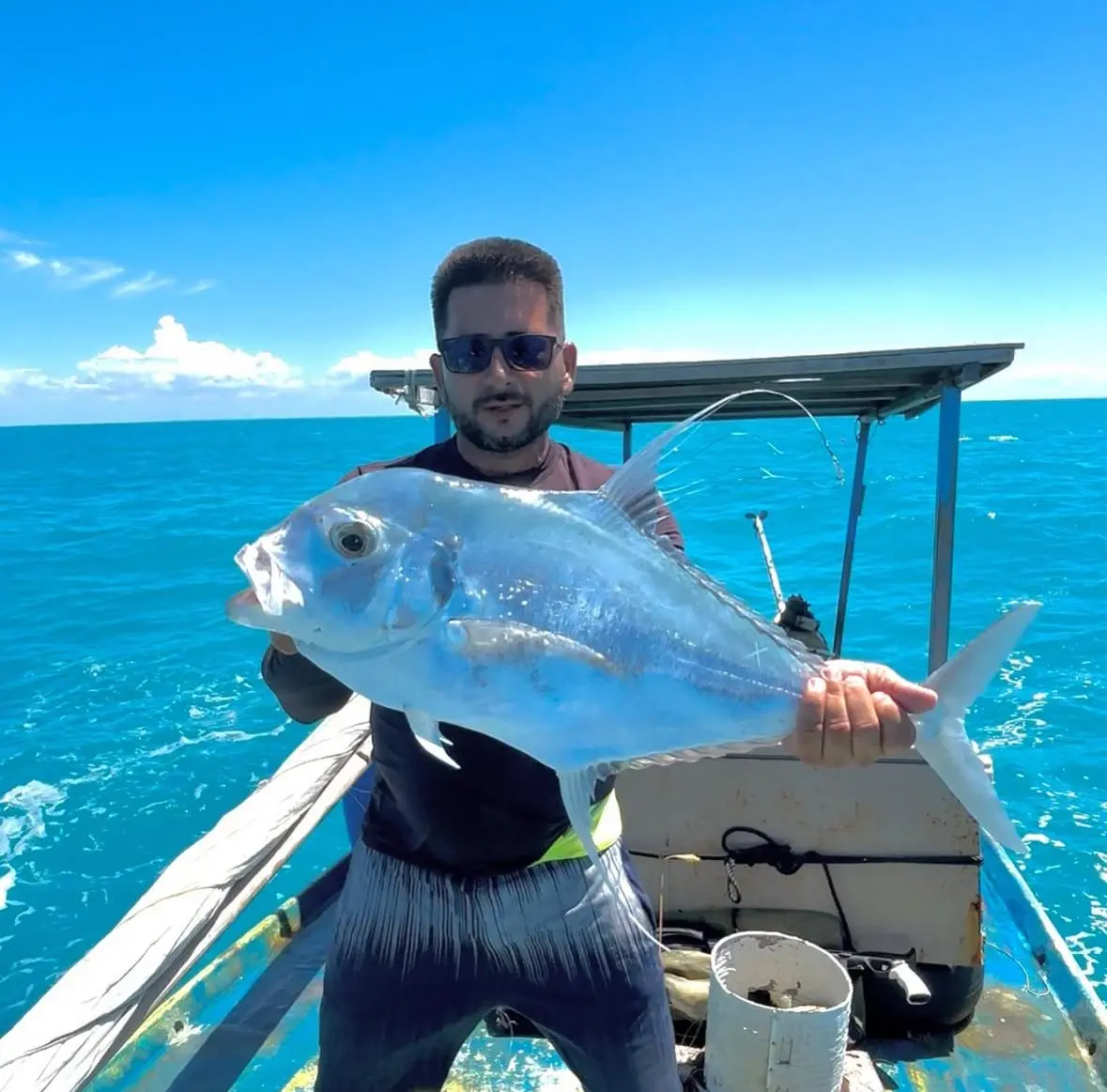 Homem branco de cabelos curtos pretos usando uma blusa fina de mangas longas e preta com uma bermuda escura. Ele segura um grande peixe azul e está em um barco em alto mar, com fundo azul do céu e do oceano.