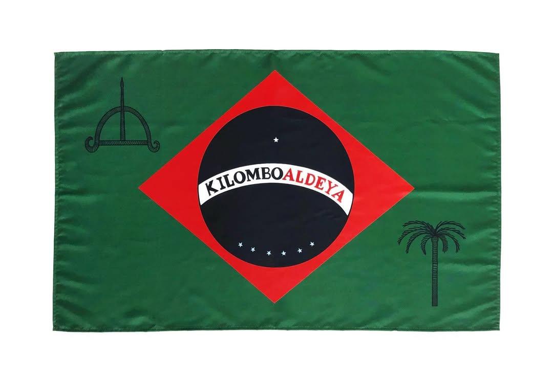 Na imagem, uma bandeira retangular, predominantemente verde-escura. No centro, há um losango vermelho que contém um círculo preto. A borda superior do círculo preto tem um semicírculo branco com o texto em vermelho 