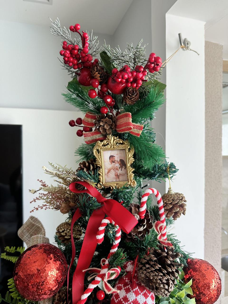 Um close detalhado de uma árvore de Natal ricamente decorada com um estilo tradicional. A árvore é adornada com folhagens verdes, galhos cobertos de neve falsa, pinhas naturais e artificiais, e cachos de frutos vermelhos brilhantes. Várias fitas e laços em tons de vermelho e padrão xadrez complementam a decoração. No centro, um pequeno porta-retrato dourado de estilo vintage exibe uma foto de um casal com um bebe no colo. Outros enfeites incluem bengalas doces vermelhas e brancas, bolas vermelhas brilhantes e um enfeite de tricô com padrão natalino na base.