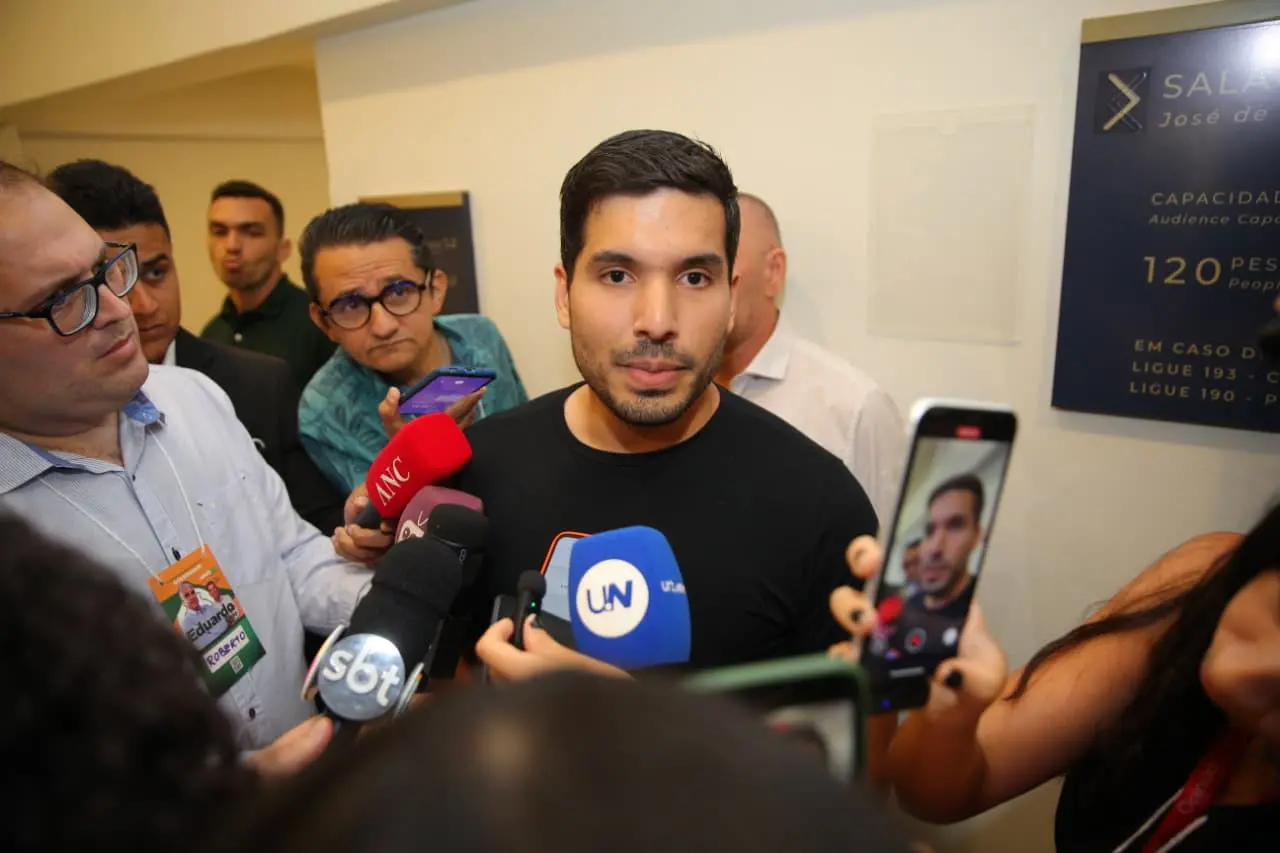 André Fernandes de camiseta preta concede entrevista em um corredor, cercado por vários jornalistas que seguram microfones e celulares. Outras pessoas observam ao fundo.