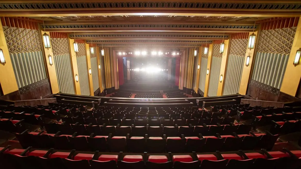 Na imagem, vista panorâmica da plateia e do palco do Cineteatro São Luiz. O interior é ricamente decorado em estilo Art Déco com poltronas de veludo vermelho e detalhes arquitetônicos dourados e verde-água nas paredes, emoldurados por um teto trabalhado. Luzes fortes e centralizadas emanam do palco, realçando o contraste escuro da plateia.