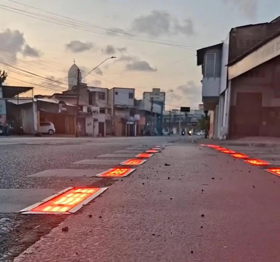 Imagem de rua com calçada iluminada por fitas de LED laranja e casas ao fundo, durante o entardecer, em uma área urbana.