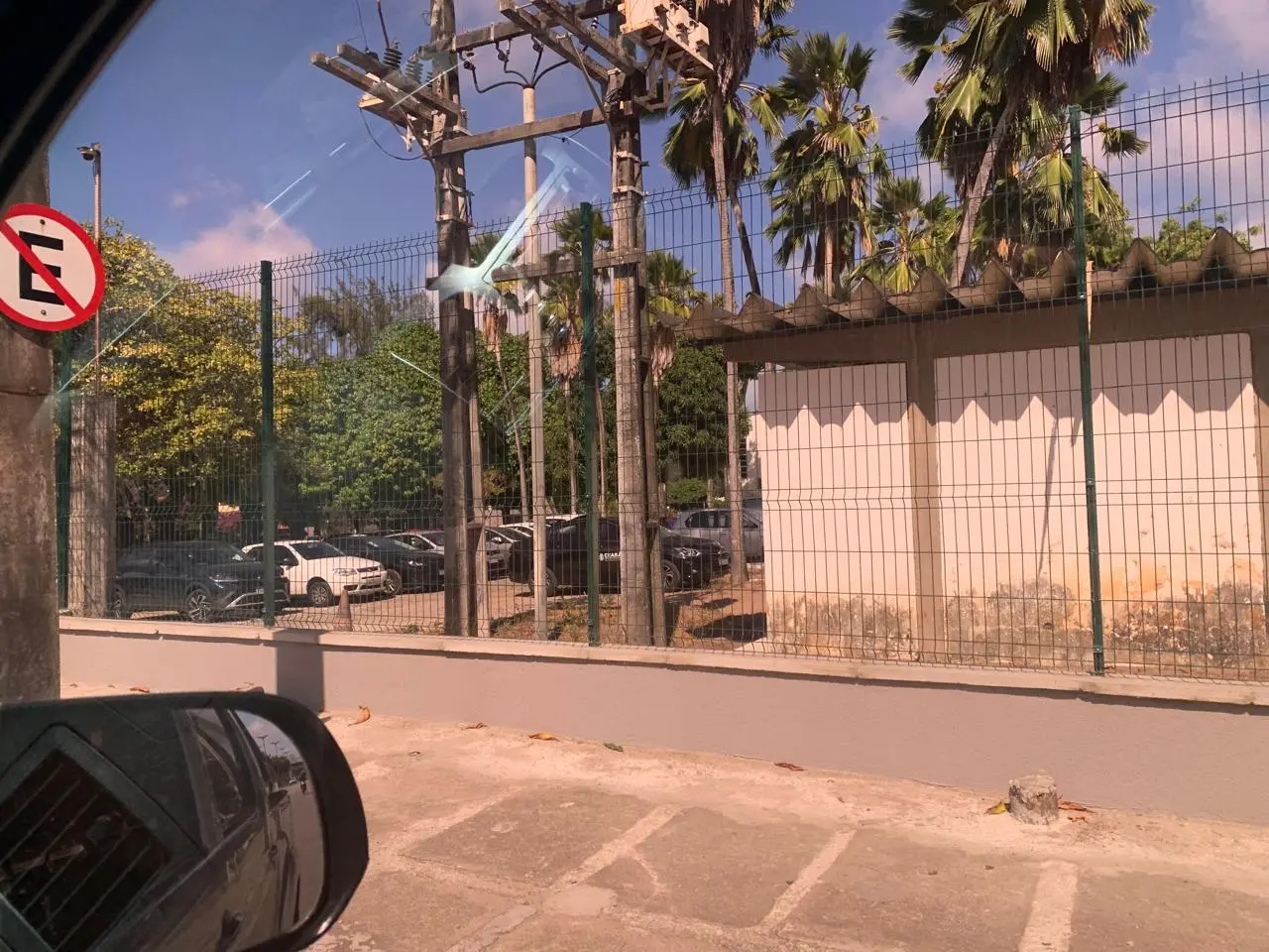 Na imagem, uma fotografia tirada de dentro de um veículo, mostrando o canto do espelho retrovisor lateral na parte inferior esquerda. A vista é de um estacionamento cercado e uma subestação elétrica simples. No primeiro plano, há um calçamento cinza com marcas de estacionamento esmaecidas e uma pequena mureta. À esquerda, um sinal de trânsito redondo de 