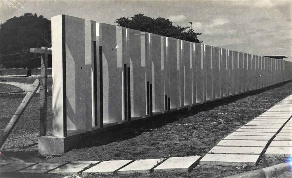 Na imagem, uma fotografia em preto e branco que mostra um longo muro de concreto com um forte apelo de arte cinética, estendendo-se em perspectiva da esquerda para a direita. O muro é composto por uma série de painéis verticais e reentrâncias, com algumas das reentrâncias preenchidas por barras verticais escuras (pretas), criando um efeito visual que muda conforme o ângulo do observador. No primeiro plano, à direita, há um caminho de lajes de concreto quadradas dispostas sobre o chão de terra. À esquerda, a paisagem é delimitada por uma cerca de arame farpado e postes rústicos, com árvores de folhagem densa ao fundo. A foto foi creditada a Sérvulo Esmeraldo.