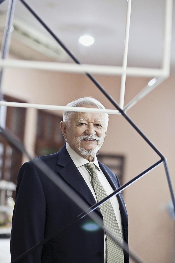 Na imagem, um retrato vertical do artista brasileiro Sérvulo Esmeraldo, tirado em 17 de abril de 2015, com crédito de Jarbas Oliveira. Ele é um homem idoso, de pele clara, com cabelos brancos e um bigode e barba brancos bem cuidados. Ele sorri levemente para a câmera. Veste um blazer marinho (azul escuro) sobre uma camisa clara e uma gravata estampada em tons de verde-oliva e bege. A composição é notável: ele está enquadrado por uma de suas esculturas geométricas de arame, que consiste em finas hastes pretas e brancas interligadas. O fundo está desfocado e mostra um interior com paredes em tons pastéis (pêssego ou rosa claro) e o início de uma escada. A iluminação é suave e focada no rosto do artista.