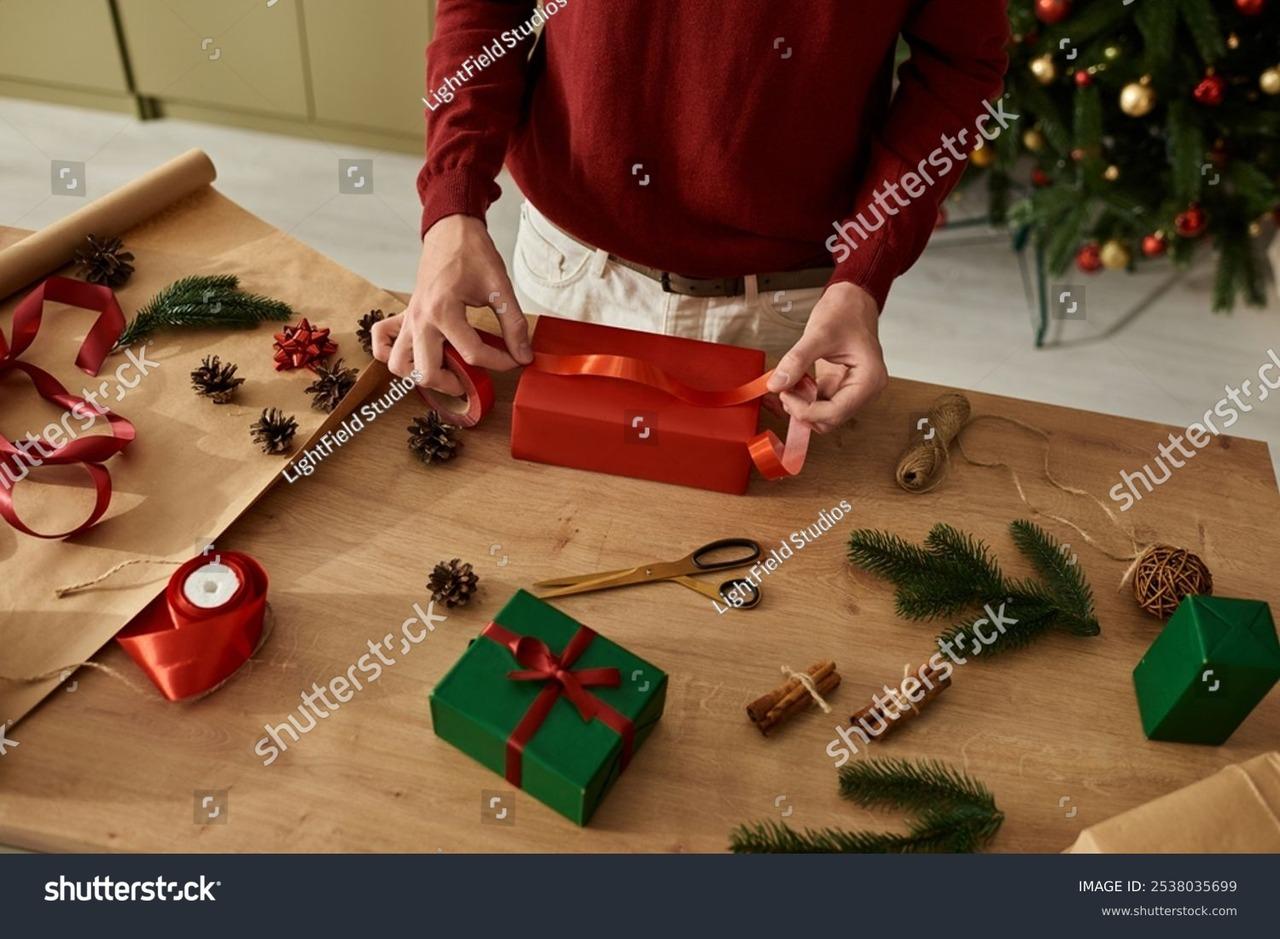 Material de embrulho, com caixa vermelha, fita vermelha, tesoura, caixa verde embrulahda e outros itens vermelhos e verdes espalhados em uma mesa de madeira. Atrás, há o torso de uma pessoa com um moletom vermelho e calça branca que está embrulhando um presente.