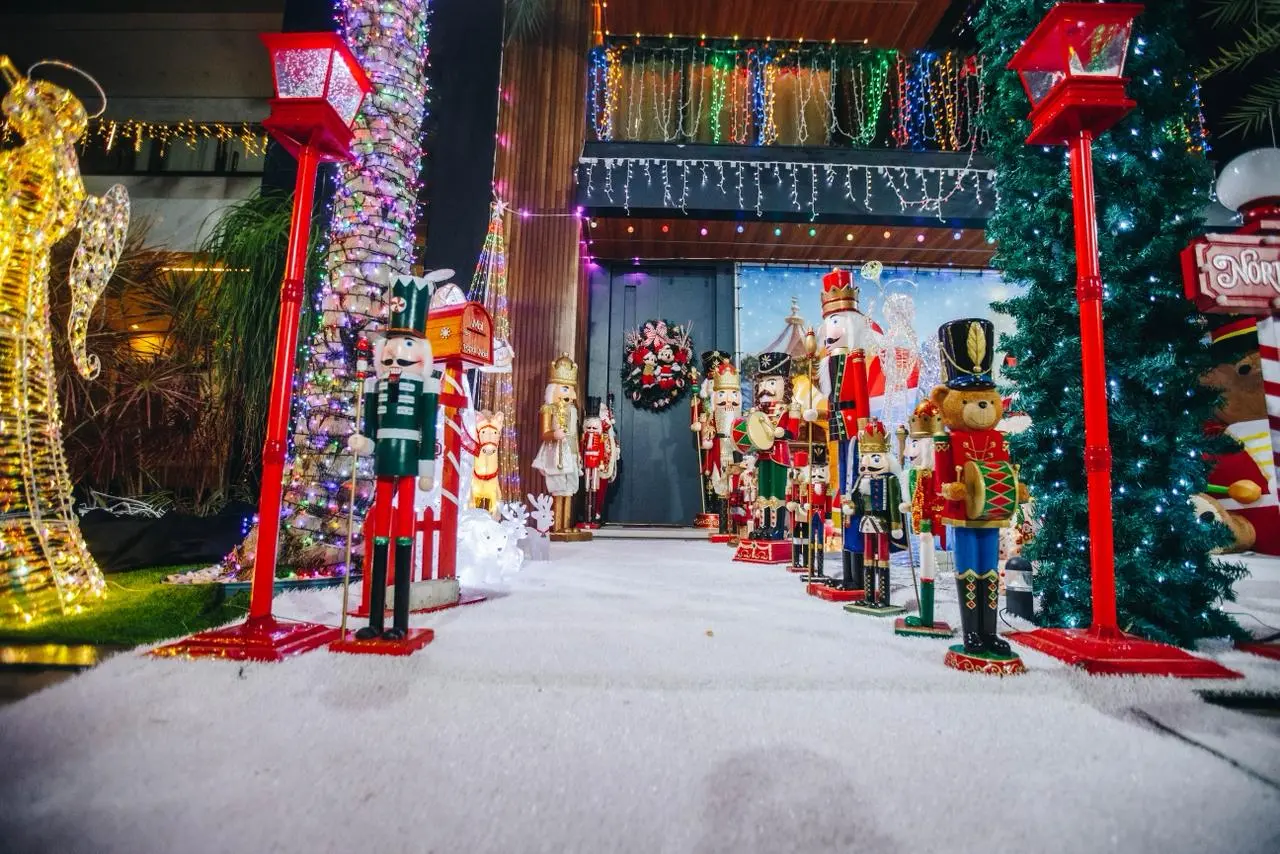 Entrada de uma casa luxuosamente decorada para o Natal, com um caminho coberto por neve artificial ladeado por dezenas de grandes soldadinhos Quebra-Nozes (Nutcrackers) e dois postes de luz vermelhos. A fachada superior está coberta de luzes coloridas e há uma grande escultura dourada iluminada de um anjo à esquerda. A porta central exibe uma coroa e a decoração evoca um tema de Polo Norte.