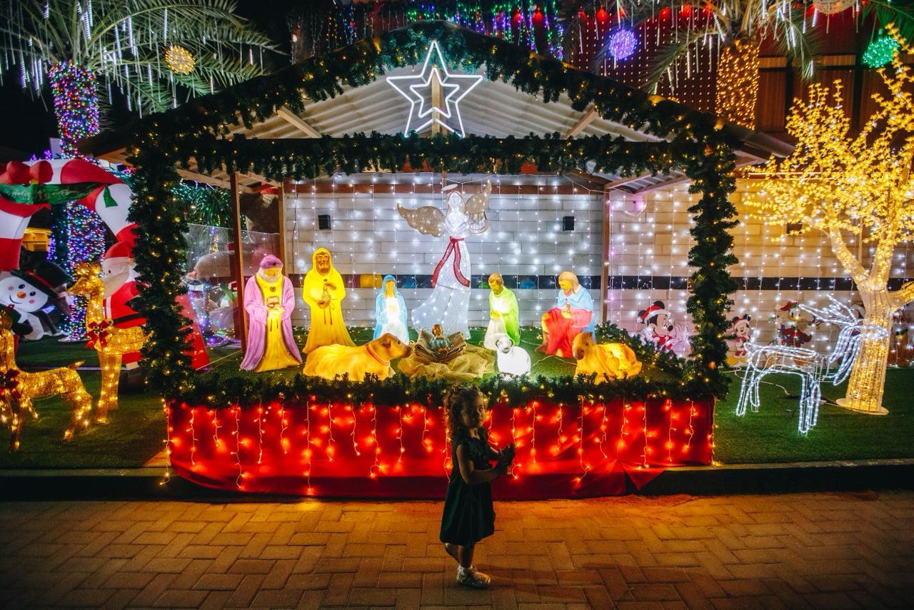 Entrada de uma casa luxuosamente decorada para o Natal, com um caminho coberto por neve artificial ladeado por dezenas de grandes soldadinhos Quebra-Nozes (Nutcrackers) e dois postes de luz vermelhos. A fachada superior está coberta de luzes coloridas e há uma grande escultura dourada iluminada de um anjo à esquerda. A porta central exibe uma coroa e a decoração evoca um tema de Polo Norte