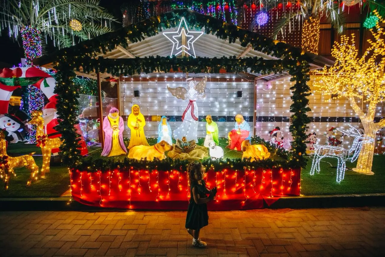 Entrada de uma casa luxuosamente decorada para o Natal, com um caminho coberto por neve artificial ladeado por dezenas de grandes soldadinhos Quebra-Nozes (Nutcrackers) e dois postes de luz vermelhos. A fachada superior está coberta de luzes coloridas e há uma grande escultura dourada iluminada de um anjo à esquerda. A porta central exibe uma coroa e a decoração evoca um tema de Polo Norte