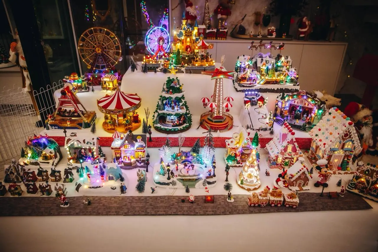 Um cenário elaborado de uma vila de Natal em miniatura, coberta de neve artificial e exibida em múltiplas camadas. O cenário altamente detalhado inclui dezenas de edifícios e casas iluminadas internamente, uma grande casa de gengibre decorada, e elementos de parque de diversões, como uma Roda Gigante e um carrossel. Pequenas figuras e árvores de Natal decoradas completam a cena festiva e colorida.