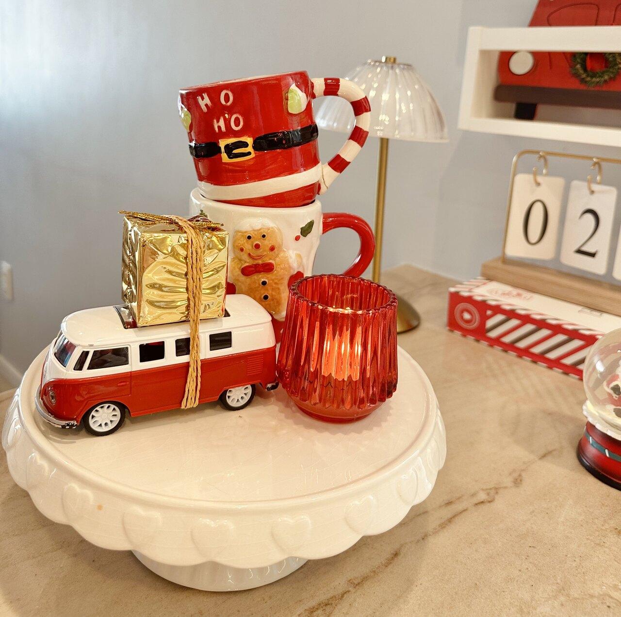 Decoração natalina, com mini ônibus branco e vermelho, pote vermelho, canecas de boneco de biscoito e de papai noel e quadradinhos de presente dourados.