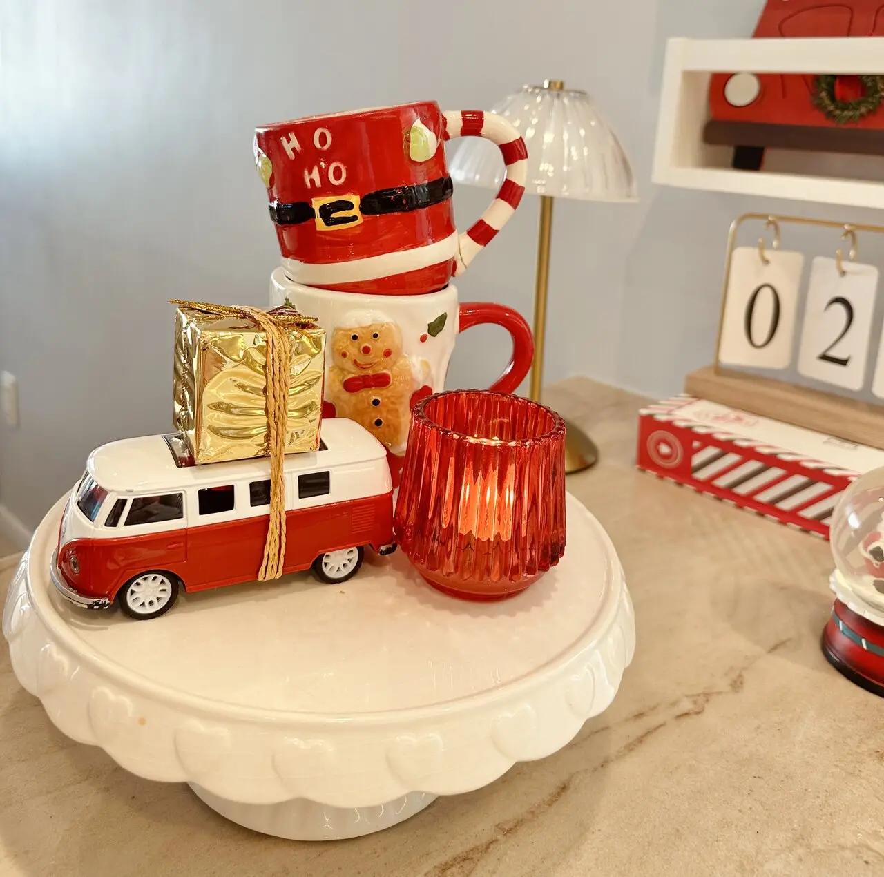 Decoração natalina, com mini ônibus branco e vermelho, pote vermelho, canecas de boneco de biscoito e de papai noel e quadradinhos de presente dourados.