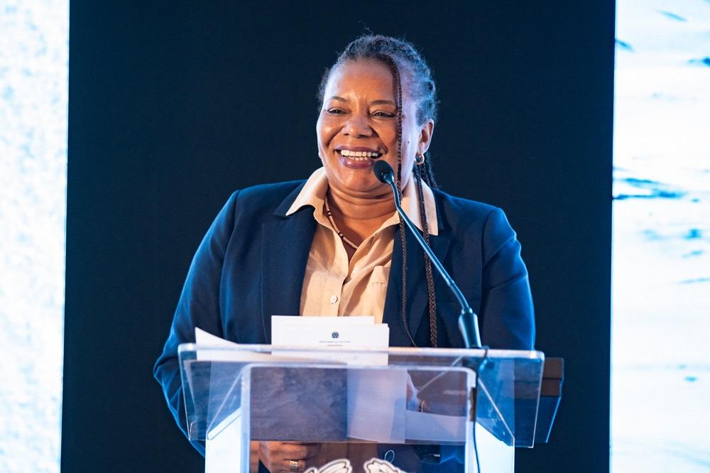 Na imagem, uma mulher de pele escura e meia-idade, com tranças escuras, sorri amplamente enquanto está atrás de um púlpito de acrílico transparente. Ela está falando em um microfone de haste fina e vestindo um blazer azul-marinho sobre uma camisa social bege ou champagne de gola aberta. No púlpito, há alguns papéis, incluindo um com um emblema impresso. A iluminação é brilhante, destacando a mulher, e o fundo é predominantemente escuro, talvez com algumas texturas de luz azuladas no canto superior direito e esquerdo, sugerindo um palco.