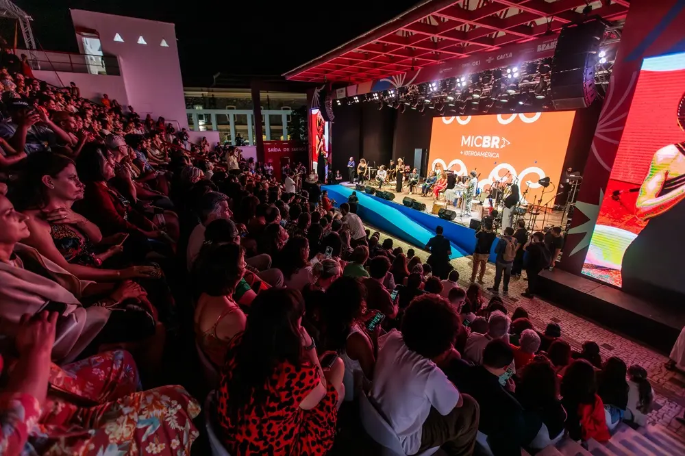 Na imagem, uma foto tirada à noite, mostrando uma grande plateia sentada em arquibancadas em um espaço ao ar livre. As pessoas estão olhando para um palco elevado, iluminado no centro. O palco tem uma tela de LED grande ao fundo, exibindo a logomarca 