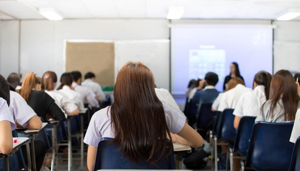 Na imagem, vista traseira de uma sala de aula ou exame com estudantes sentados em carteiras azuis, voltados para uma tela de projeção e um instrutor à frente.