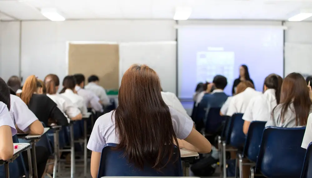Na imagem, vista traseira de uma sala de aula ou exame com estudantes sentados em carteiras azuis, voltados para uma tela de projeção e um instrutor à frente.