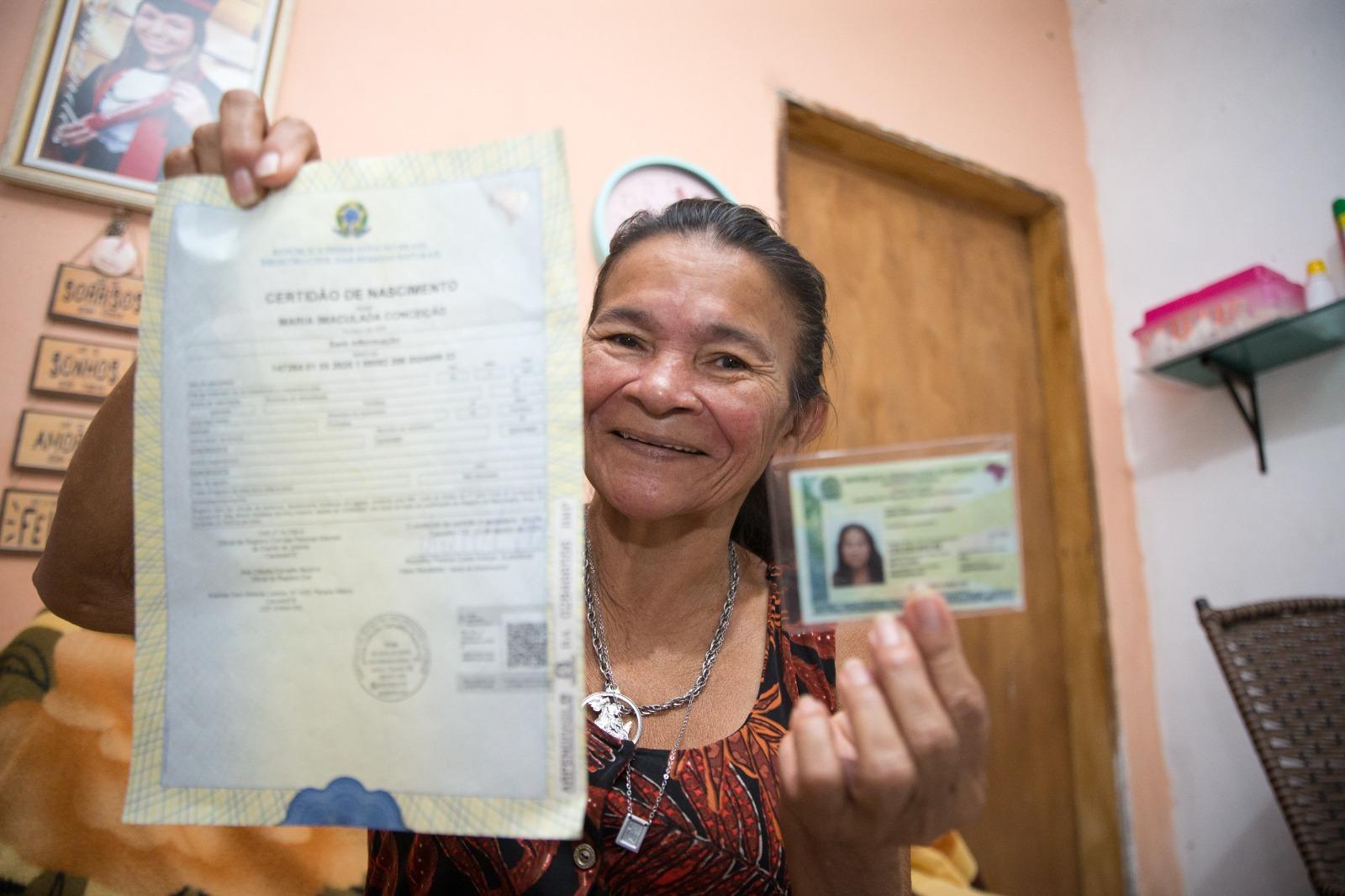 Na imagem, retrato em plano médio de uma mulher brasileira mais velha, de pele morena, sorrindo e olhando diretamente para a câmera, segurando dois documentos. Com a mão esquerda, ela segura, bem próxima e em foco suave, uma Certidão de Nascimento (Certidão de Nascimento) brasileira. Com a mão direita, em foco mais nítido, ela segura uma carteira de identidade ou documento de registro nacional (RG) plastificado, que mostra sua foto. Ela está vestindo uma blusa estampada e um colar. O fundo mostra uma parede pêssego/rosa e uma porta de madeira.