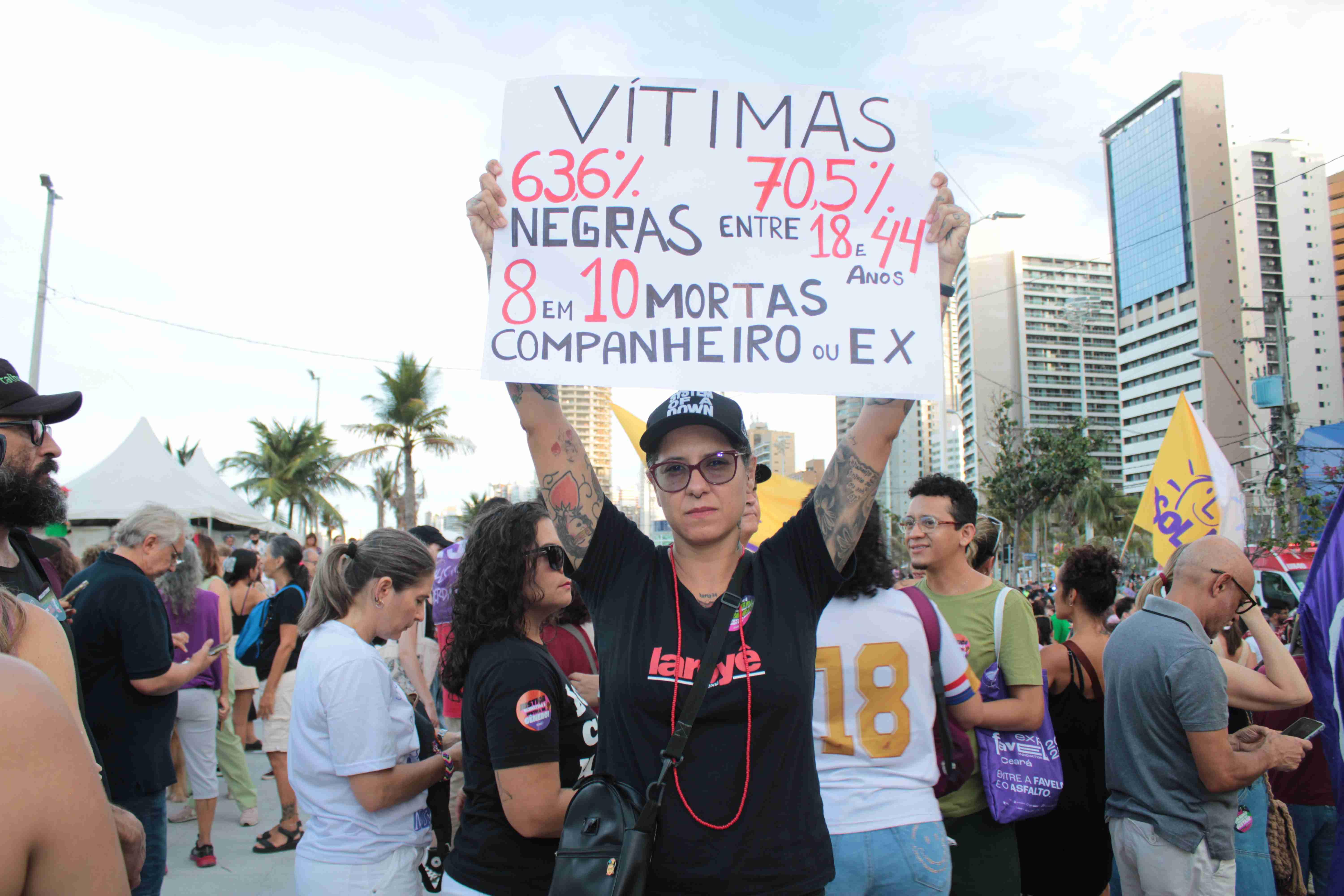Imagem de um protesto mostra uma mulher segurando um cartaz branco com letras grandes vermelhas e pretas, detalhando estatísticas sobre feminicídio, com foco em vítimas negras e jovens e a autoria dos crimes por parceiros. Edifícios e uma multidão são visíveis ao fundo.
