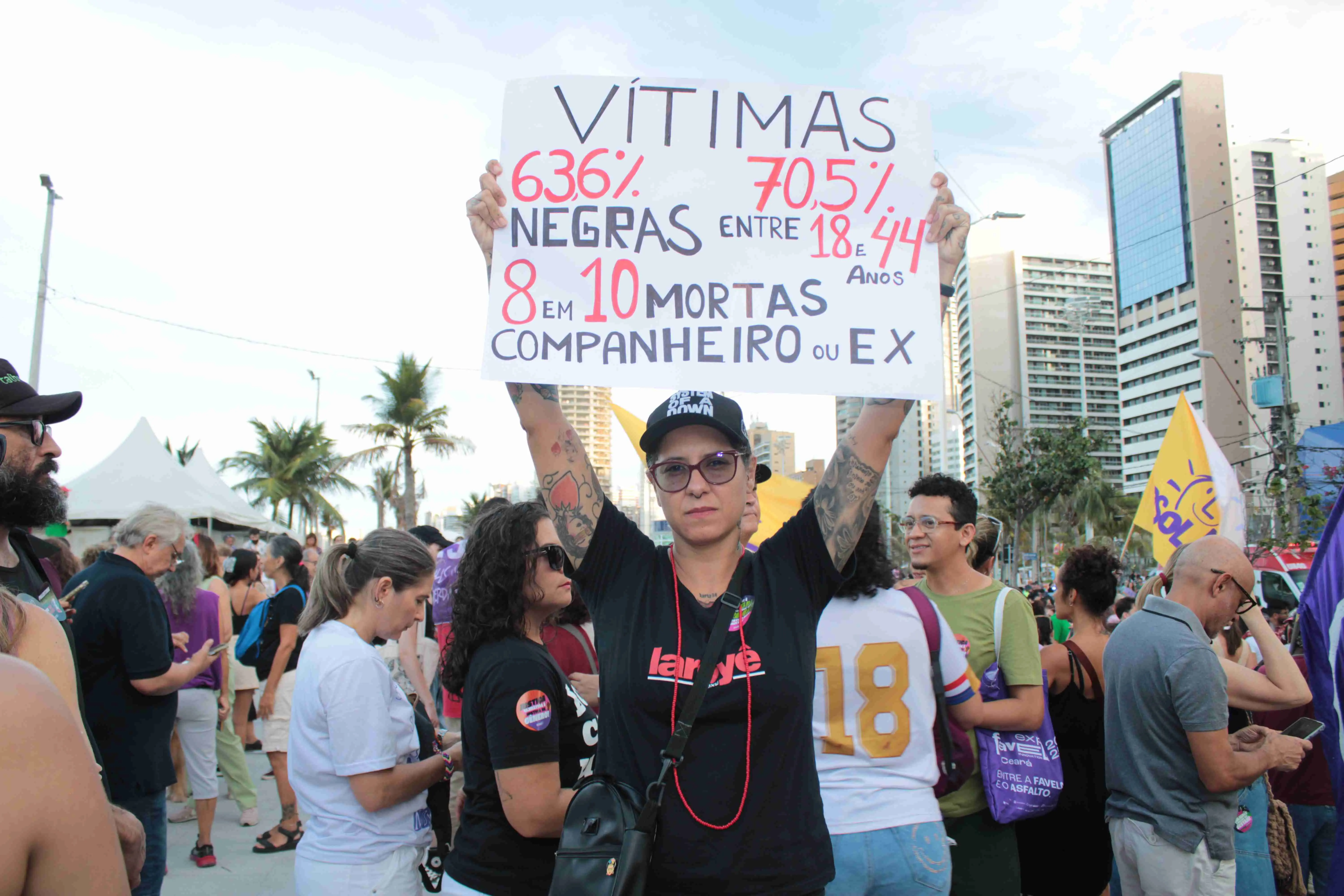 Imagem de um protesto mostra uma mulher segurando um cartaz branco com letras grandes vermelhas e pretas, detalhando estatísticas sobre feminicídio, com foco em vítimas negras e jovens e a autoria dos crimes por parceiros. Edifícios e uma multidão são visíveis ao fundo.