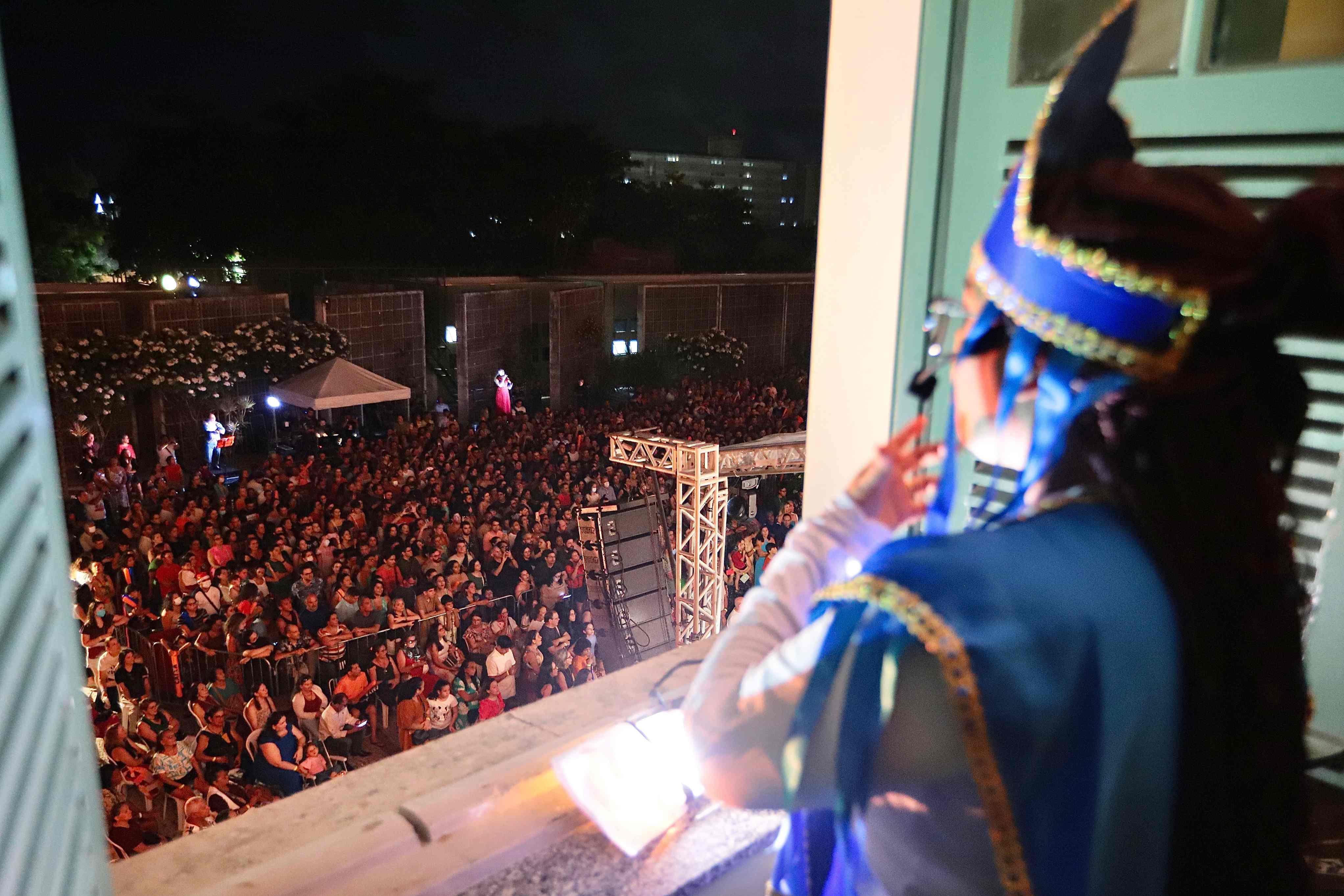 A foto noturna é tirada da perspectiva de uma janela, onde uma pessoa em primeiro plano, vista de costas e vestindo uma elaborada fantasia azul e dourada com um chapéu alto, observa a cena. Abaixo, uma multidão densa está reunida em um pátio escuro para assistir a um evento, com a luz focando na área do público e estruturas de palco visíveis à frente. Ao fundo, no meio da plateia, outra figura em roupa vermelha pode ser vista se apresentando sob as luzes. A imagem captura o momento da apresentação vista de cima, a partir do ponto de vista do artista na janela.