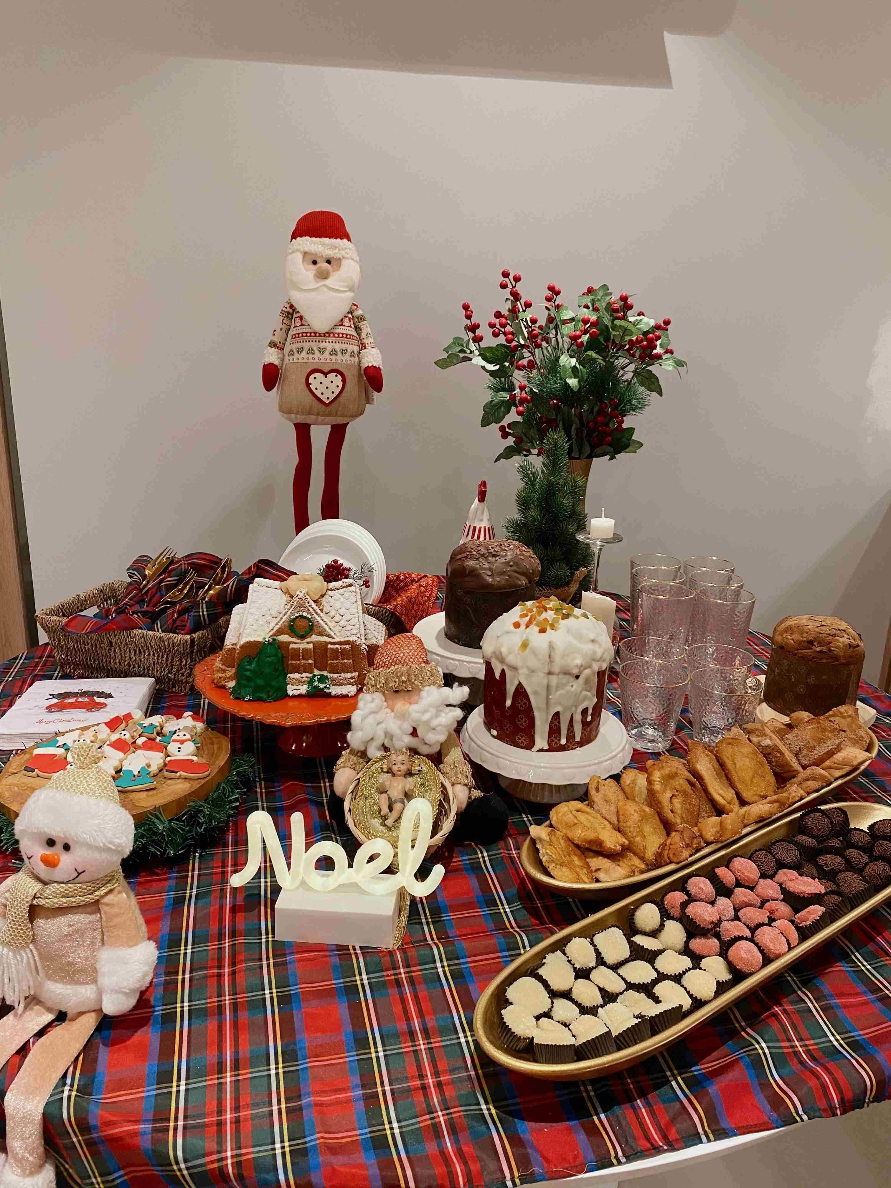 Esta mesa de celebração apresenta uma composição rica e festiva, utilizando uma toalha xadrez em tons de vermelho, verde e preto como base para um banquete natalino repleto de doces e decorações temáticas. O arranjo central destaca-se com uma variedade de panetones e bolos, incluindo um generosamente coberto com glacê branco, e é ladeado por elementos clássicos como uma elaborada casa de gengibre e uma travessa longa que exibe docinhos coloridos (brigadeiros e trufas) junto a salgados de massa folhada. Figuras natalinas, como um Papai Noel de pano, um boneco de neve de pelúcia e um adorno com a palavra 