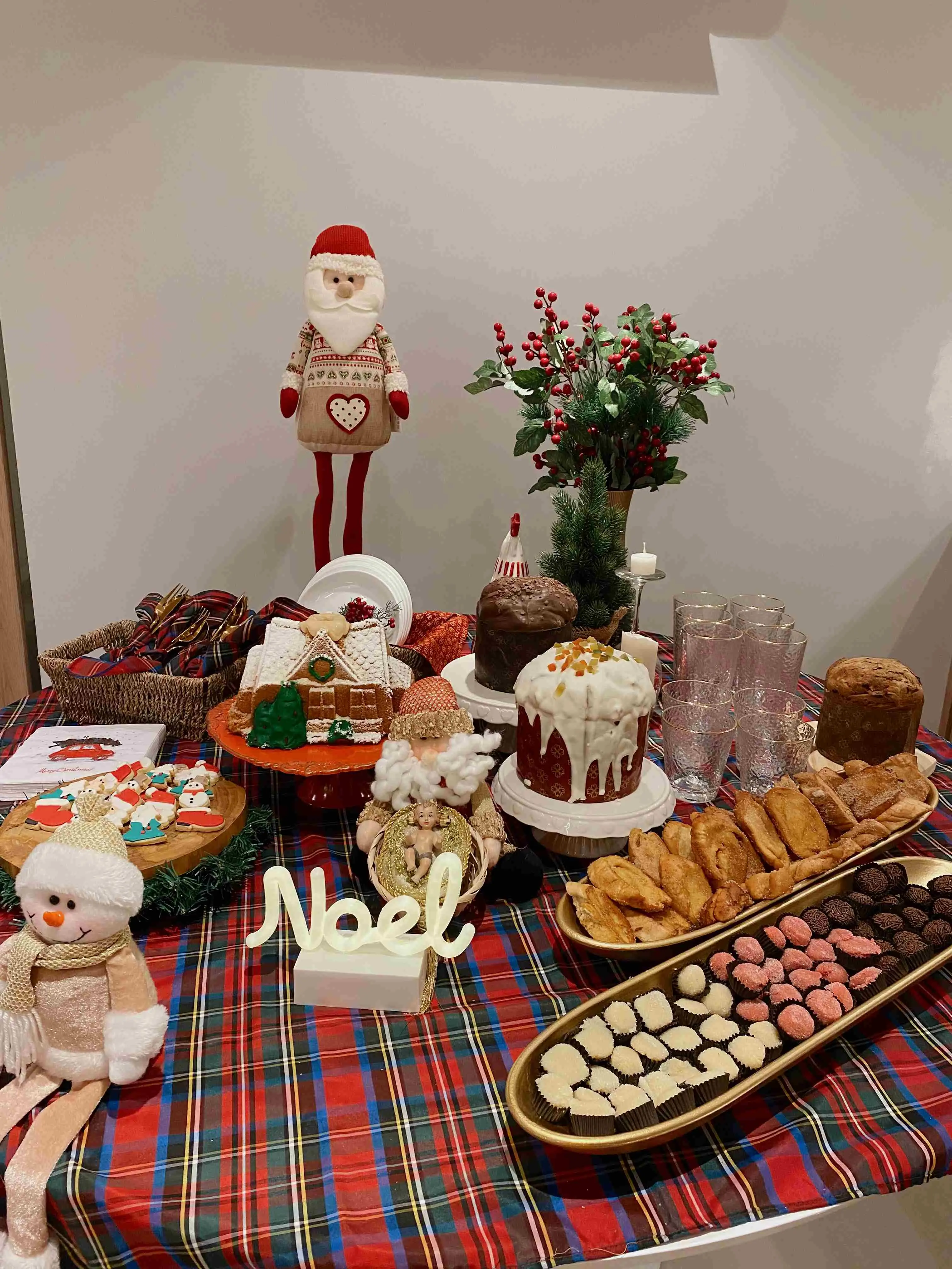 Esta mesa de celebração apresenta uma composição rica e festiva, utilizando uma toalha xadrez em tons de vermelho, verde e preto como base para um banquete natalino repleto de doces e decorações temáticas. O arranjo central destaca-se com uma variedade de panetones e bolos, incluindo um generosamente coberto com glacê branco, e é ladeado por elementos clássicos como uma elaborada casa de gengibre e uma travessa longa que exibe docinhos coloridos (brigadeiros e trufas) junto a salgados de massa folhada. Figuras natalinas, como um Papai Noel de pano, um boneco de neve de pelúcia e um adorno com a palavra 