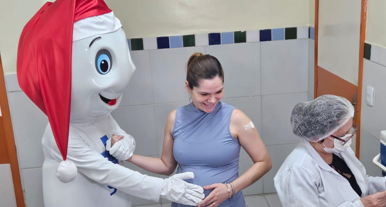 Médica sorridente recebendo a visita de um personagem de festa de Natal em hospital, vestindo uma fantasia de Papai Noel, durante campanha de saúde.