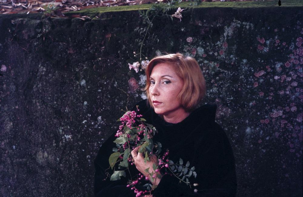 Na imagem, retrato colorido da escritora Clarice Lispector, tirado por Maureen Bisilliat. Clarice está em primeiro plano, com cabelos curtos e ruivos, vestindo uma roupa escura que contrasta com sua pele clara. Ela segura um ramo de flores com pequenas inflorescências cor-de-rosa, que cobre parte de seu colo e ombro. Ao fundo, há um muro de pedra coberto por musgo escuro e manchas rosa-claro ou lilás. A expressão da escritora é séria e introspectiva.