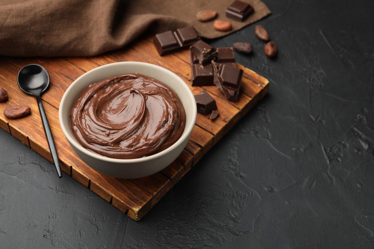 Pote branco com mousse de chocolate. Ele está posicionado em cima de uma tábua de madeira, com pedaços de chocolate e uma colhe larga aos lados.