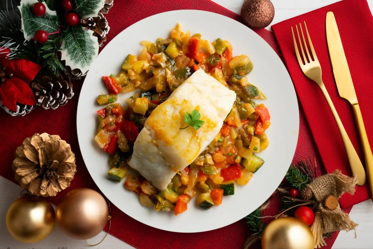 Foto de cima de um prato com bacalhau preparado e legumes ao redor. Ao lado esquerdo, alguns enfeites natalinos e, ao direito, talheres dourados.