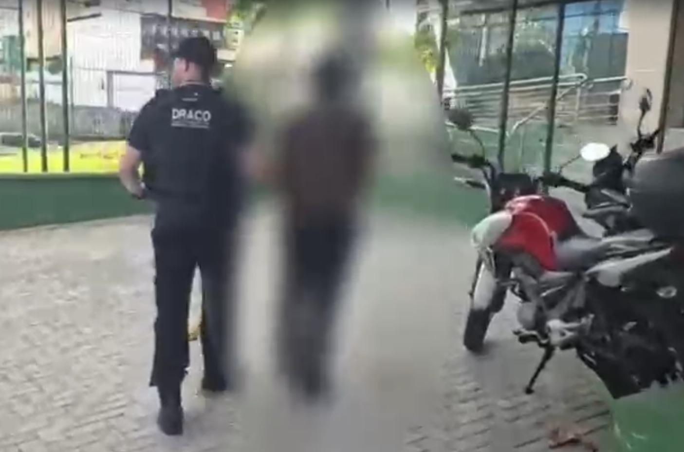 A foto mostra um homem sendo conduzido por um policial civil, fardado e com colete escrito Draco, em Fortaleza.