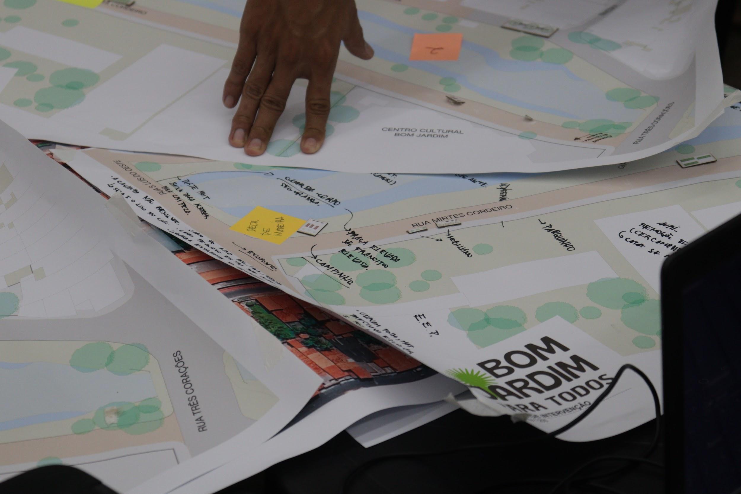 Mão sobre mapas urbanos com anotações e post-its em uma mesa.