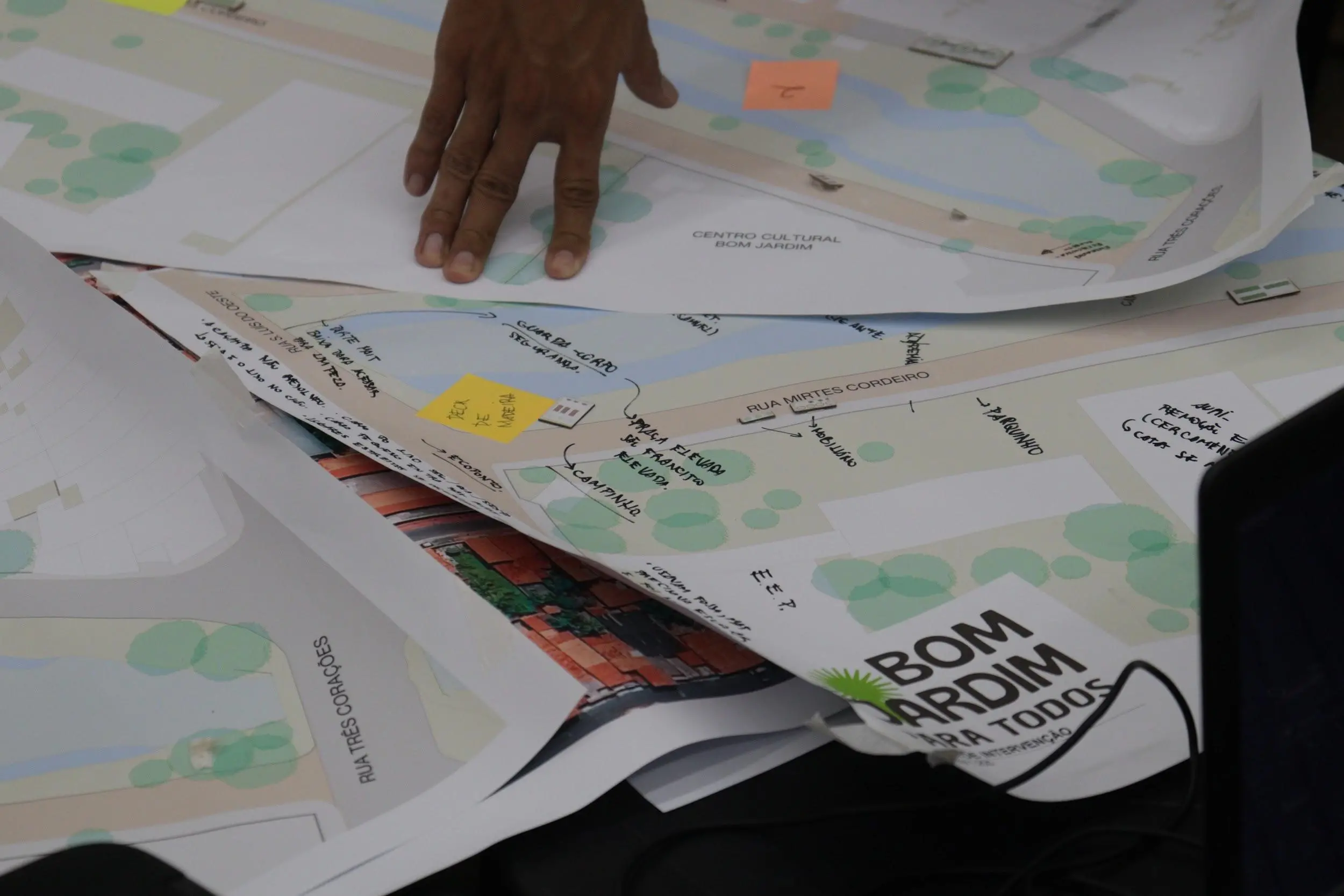 Mão sobre mapas urbanos com anotações e post-its em uma mesa.