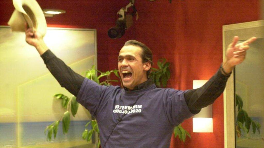 Participante do Big Brother Brasil comemorando com os braços erguidos, segurando um chapéu em uma das mãos, usando camiseta com texto em ambiente interno decorado com plantas e paredes vermelhas.