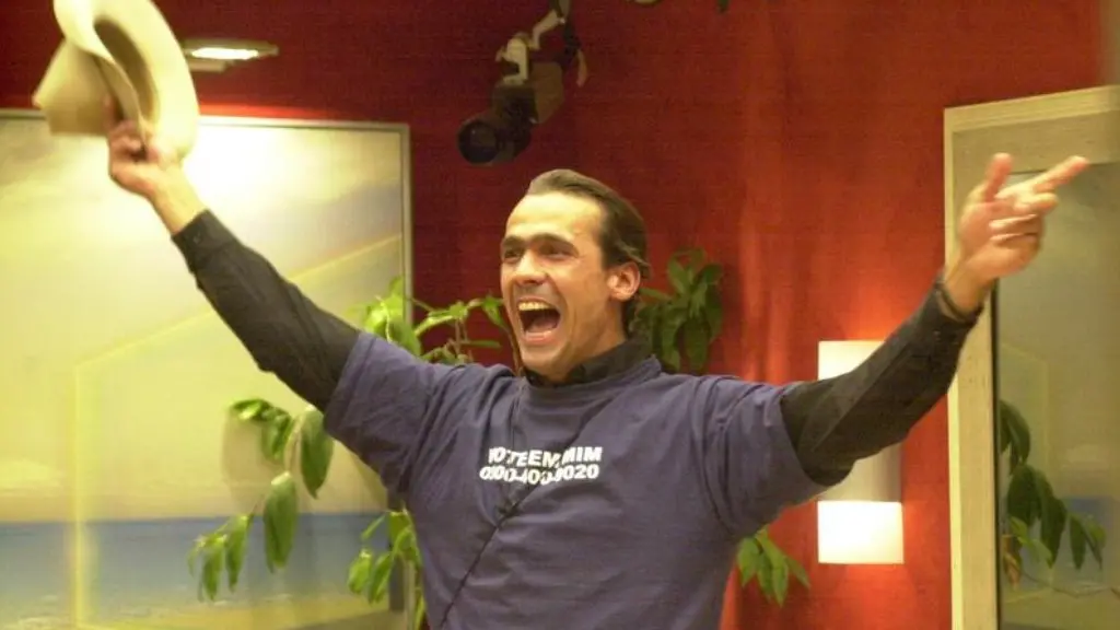 Participante do Big Brother Brasil comemorando com os braços erguidos, segurando um chapéu em uma das mãos, usando camiseta com texto em ambiente interno decorado com plantas e paredes vermelhas.