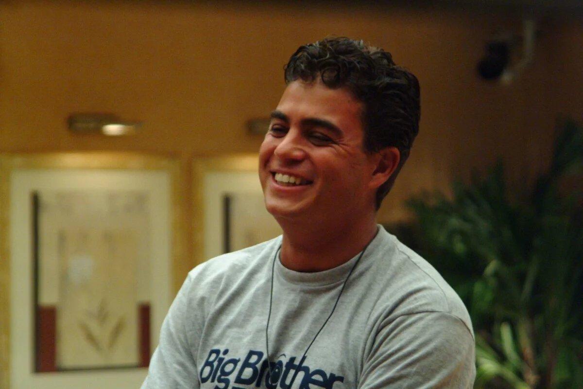 Participante do Big Brother Brasil sorrindo vestindo camiseta cinza com logo do programa, em ambiente interno com paredes claras, quadros decorativos e plantas ao fundo.