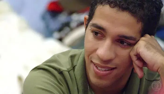 Pessoa vestindo camiseta verde com gola polo, apoiando a cabeça na mão, em ambiente interno com fundo desfocado e logotipo da TV Globo no canto inferior direito.