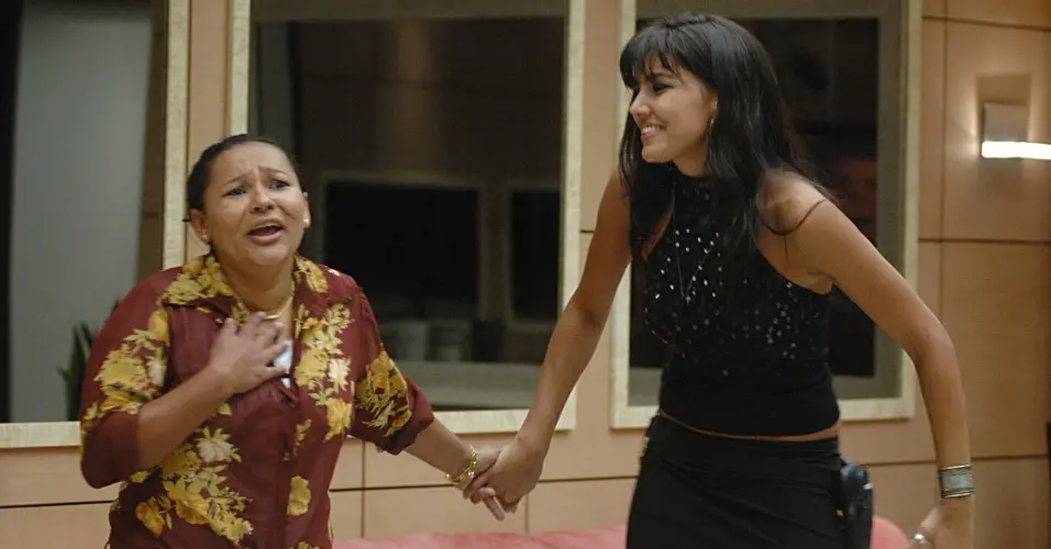 Pessoa usando camisa estampada com flores em tons de amarelo e vermelho, posicionada em ambiente interno com paredes claras e janelas ao fundo, segurando a mão de outra pessoa parcialmente visível.