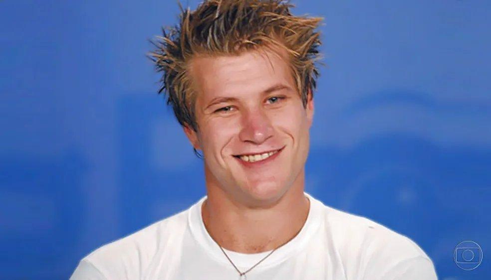 Pessoa com cabelo estilizado em pontas, vestindo camiseta branca simples e colar com pingente, posicionada diante de fundo azul sólido com logotipo da TV Globo no canto inferior direito.