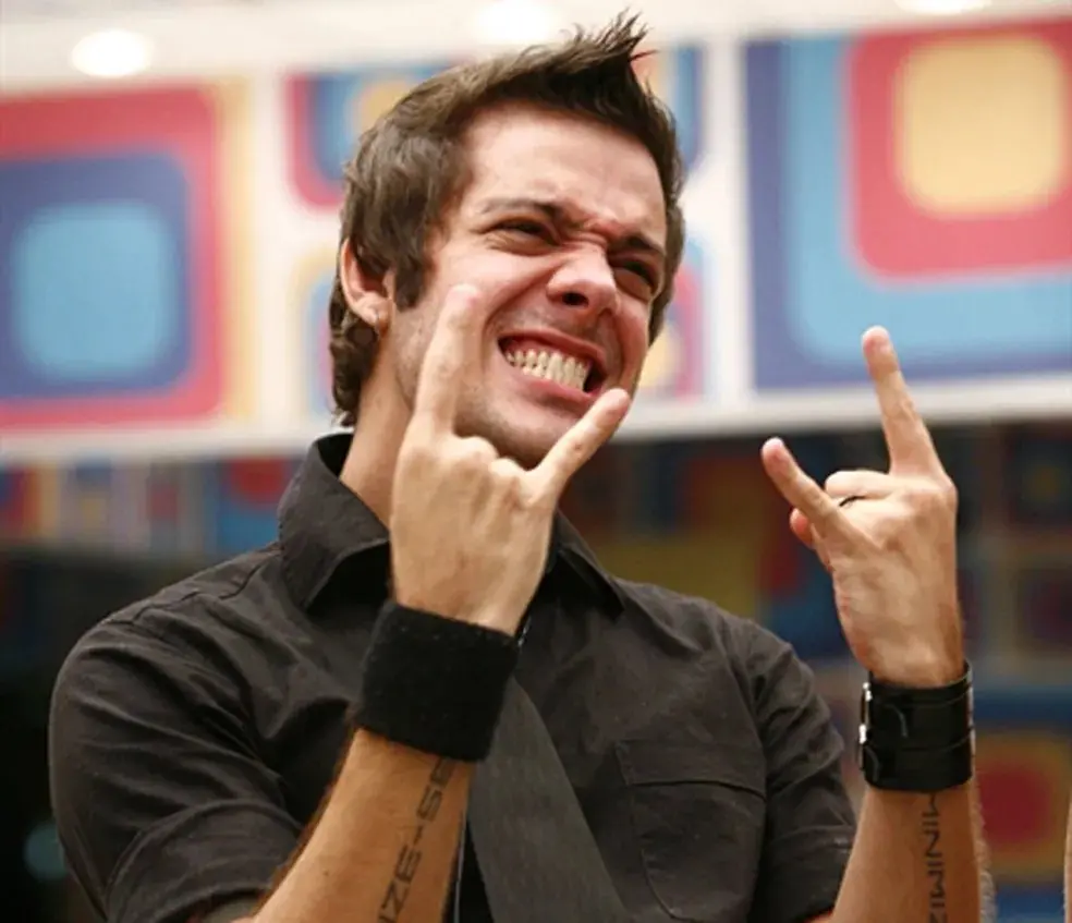 Pessoa vestindo camisa preta com pulseiras largas nos punhos e tatuagens visíveis nos antebraços, fazendo gesto de mãos com sinal de rock, em ambiente colorido com padrões geométricos ao fundo.