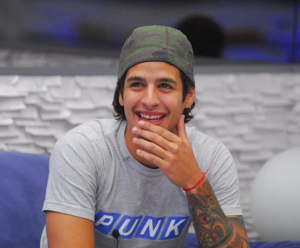 Pessoa usando camiseta cinza com a palavra 'PUNK' em destaque, boné camuflado e pulseira vermelha, exibindo tatuagem colorida no braço, sentada em ambiente interno com parede texturizada branca e sofá azul.