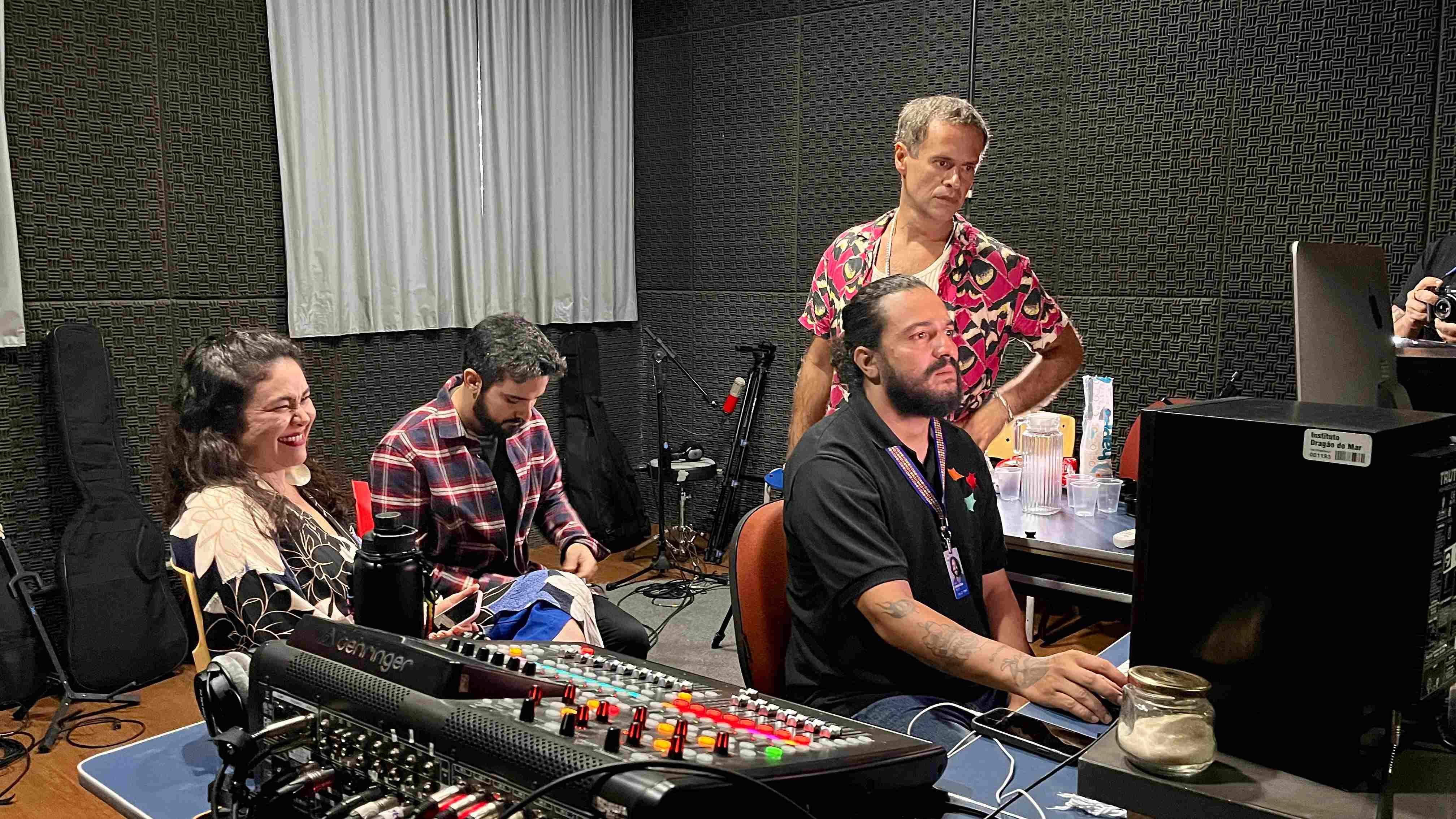 Em um ambiente de estúdio de gravação profissional, com paredes forradas em espuma acústica preta, quatro pessoas interagem em torno de uma grande mesa de som Behringer que domina o primeiro plano. Sentada à esquerda, uma mulher de blusa floral estampada sorri, olhando para fora da cena, enquanto um homem ao centro, de camisa xadrez em tons de vinho, está inclinado e concentrado em algo no colo. À direita, um homem de barba, cabelos longos e camisa polo preta está sentado e focado na mesa de som e no computador, manipulando os equipamentos. Atrás dele, em pé, está Marcelo Jeneci – vestindo sua camisa de estampa vibrante e colorida –, que observa a cena com as mãos na cintura e uma expressão séria, sugerindo um momento de tutoria ou ajustes técnicos do grupo.